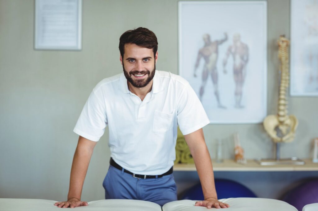 male chiropractor standing in his clinic leaning on massage table